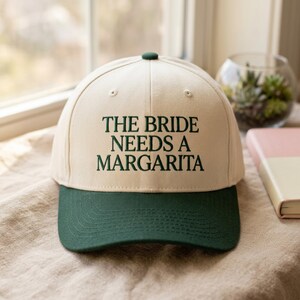 Może przedstawiać: Kremowa czapka z daszkiem z ciemnozielonym daszkiem i guzikiem. Tekst "THE BRIDE NEEDS A MARGARITA" jest wyhaftowany na przodzie ciemnozielonym kolorem. Zabawny dodatek na wieczór panieński lub imprezę ślubną.