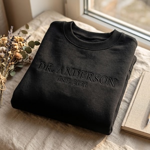 Puede incluir: Sudadera negra de cuello redondo con texto bordado "DR. ANDERSON EST. 2026". La sudadera está doblada sobre una superficie beige, junto a un libro y un ramo de flores secas. La imagen está iluminada por luz natural.