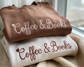 May include: Two sweatshirts, one brown and one white, are stacked. The brown sweatshirt has white embroidered text that reads "Coffee & Books." The white sweatshirt has brown embroidered text that reads "Coffee & Books."