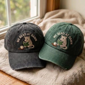 May include: Two baseball caps, one charcoal gray and one forest green, each featuring an embroidered graphic of an opossum reading a green book with a small cup of coffee nearby and the text "WE READ AT DAWN" arched above the illustration.