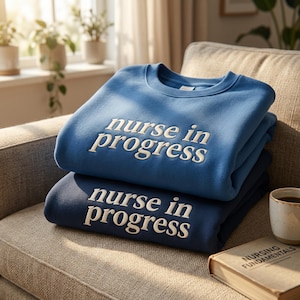 Pode incluir: Duas camisolas de gola redonda dobradas em tons de azul, empilhadas num sofá. A camisola de cima é azul-média e a de baixo é azul-marinho escuro. Ambas apresentam o texto bordado a branco "nurse in progress" na zona do peito.