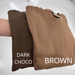 May include: Two crewneck sweatshirts featuring a solid color design. One sweatshirt is a deep dark chocolate brown, while the other is a lighter tan brown. The text DARK CHOCO is printed on the darker garment and BROWN on the lighter one.