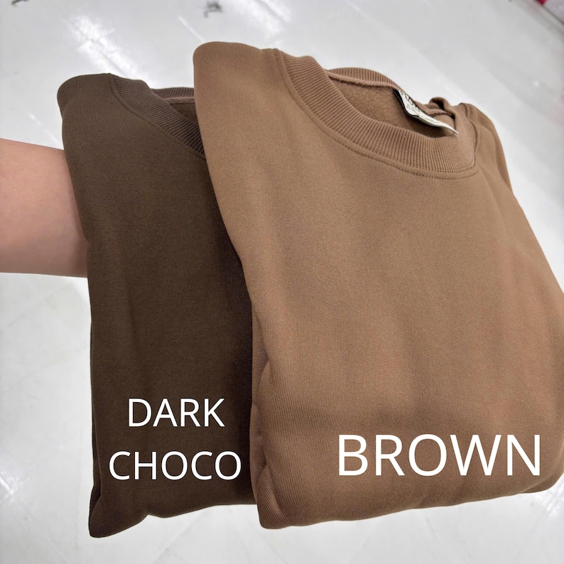 May include: Two crewneck sweatshirts featuring a solid color design. One sweatshirt is a deep dark chocolate brown, while the other is a lighter tan brown. The text DARK CHOCO is printed on the darker garment and BROWN on the lighter one.