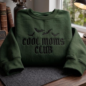 Puede incluir: Una sudadera de cuello redondo color verde bosque con texto bordado en negro que dice "cool moms CLUB" en una fuente de estilo gótico, situada debajo de cuatro pequeñas siluetas de murciélagos voladores bordadas en negro en el pecho.