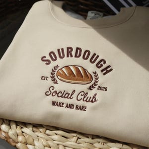 Può includere: Felpa beige con ricamo "Sourdough Social Club". Il design include una pagnotta, foglie di alloro e la scritta "Wake and Bake". La felpa è piegata su un cestino intrecciato.