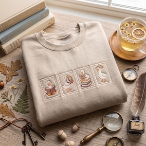 May include: A beige crewneck sweatshirt featuring four embroidered rectangular patches, each depicting a white goose in different poses with books. The geese are shown reading, carrying a stack of books, sitting on books, and balancing a book on its head.