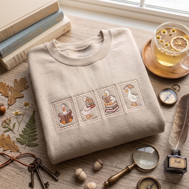 May include: A beige crewneck sweatshirt featuring four embroidered rectangular patches, each depicting a white goose in different poses with books. The geese are shown reading, carrying a stack of books, sitting on books, and balancing a book on its head.