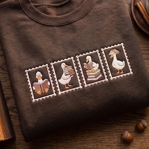 May include: A dark brown crewneck sweatshirt featuring four embroidered rectangular patches resembling postage stamps. Each patch depicts a white duck with orange feet interacting with books, including reading, carrying a stack, and balancing one on its head.