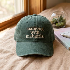 Peut inclure: Casquette de baseball verte avec le texte "mahjong with mahgirls" brodé dans une couleur plus claire. La casquette est posée sur une surface beige. Un petit terrarium en verre avec des plantes est en arrière-plan.