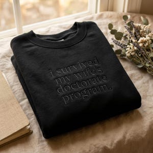 May include: Black sweatshirt with the phrase "i survived my wife's doctorate program" in embossed lettering. The sweatshirt is folded and resting on a beige surface, with a book and flowers in the background.