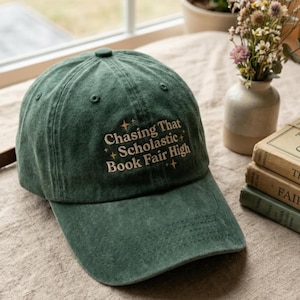 May include: A dark green baseball cap with the embroidered text "Chasing That Scholastic Book Fair High" in off-white. The cap is made of a soft material and has a curved brim. The cap is sitting on a beige surface.