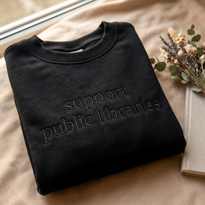 May include: Black crewneck sweatshirt with the words "support public libraries" embroidered in a tone-on-tone design. The sweatshirt is folded on a beige surface, with a window and a bouquet of dried flowers in the background.