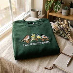 May include: A dark green sweatshirt with an embroidered design of four colorful birds perched on a branch. The text "EASILY DISTRACTED BY BIRDS" is below the birds. The sweatshirt is folded on a table with a neutral-colored cloth.