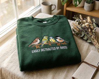 May include: A dark green sweatshirt with an embroidered design of four colorful birds perched on a branch. The text "EASILY DISTRACTED BY BIRDS" is below the birds. The sweatshirt is folded on a table with a neutral-colored cloth.