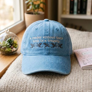 Op de afbeelding: Lichtblauwe baseballcap met geborduurde tekst: "a reader without their book is a tragedy." Onder de tekst staan geborduurde draken. De pet ligt op een beige oppervlak.