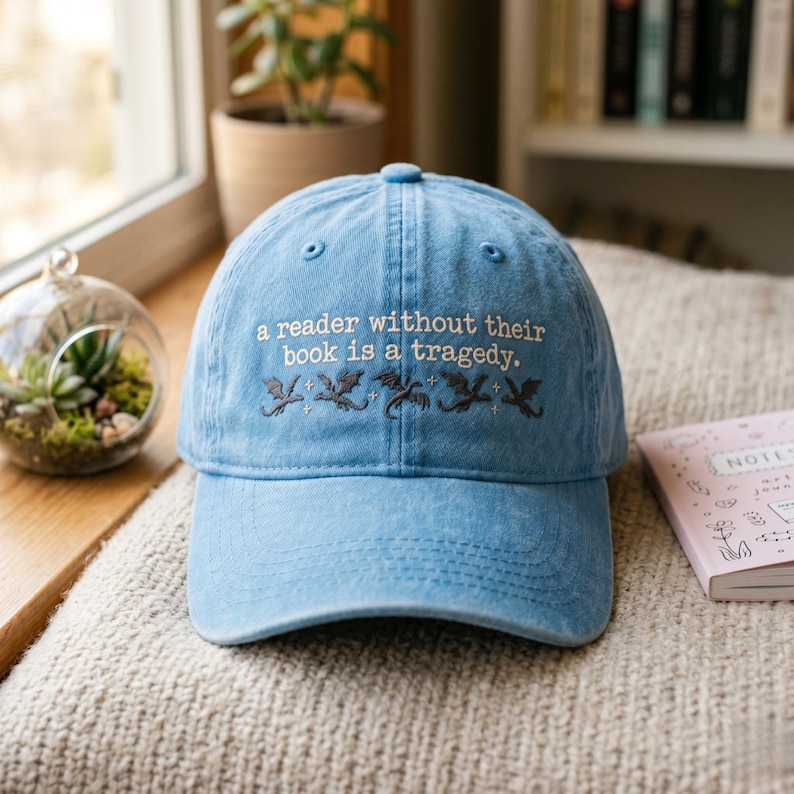 Op de afbeelding: Lichtblauwe baseballcap met geborduurde tekst: "a reader without their book is a tragedy." Onder de tekst staan geborduurde draken. De pet ligt op een beige oppervlak.