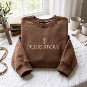 Op de afbeelding: Een bruin sweatshirt met ronde hals en een geborduurd beige kruis boven de tekst "TRUE STORY" in een schreeflettertype. Het kledingstuk heeft lange mouwen met geribbelde boorden en een geribbelde halslijn met een strak ontwerp.
