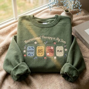 May include: Green sweatshirt with the text "Recreational Therapy is My Jam" and illustrations of jars labeled "Support", "Adapt Ability", "Motor Learning", and "Play". The jars are filled with blueberries and strawberries. The sweatshirt is folded on a beige surface.