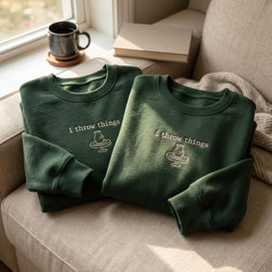 Könnte beinhalten: Zwei dunkelgrüne Sweatshirts mit Rundhalsausschnitt und weißer Stickerei mit der Aufschrift i throw things über einer Strichzeichnung einer Töpferscheibe mit einer Vase. Die Sweatshirts haben lange Ärmel sowie gerippte Bündchen.
