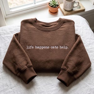 Puede incluir: Una sudadera de color marrón oscuro con cuello redondo y el texto "life happens cats help." bordado en blanco en el centro del pecho. La prenda cuenta con mangas largas con puños acanalados y un cuello acanalado de diseño sencillo.