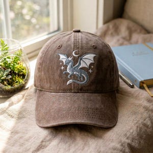 May include: A brown baseball cap featuring an embroidered design of a grey dragon with white wings, positioned beneath a crescent moon and surrounded by small four-pointed stars. The cap has a distressed, washed-out fabric texture.
