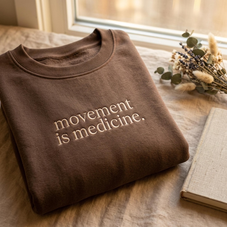 May include: A brown sweatshirt with the embroidered text "movement is medicine." The crew neck sweatshirt is made of a soft material. The sweatshirt is folded on a neutral-colored surface.
