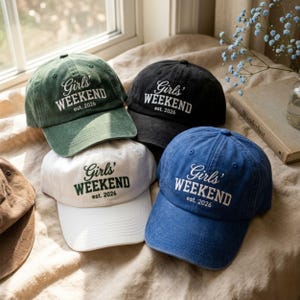 May include: Several baseball caps in shades of green, black, white, and blue. Each cap displays the text "Girls' Weekend est. 2026". The caps are placed on a textured surface, with a window and flowers in the background.
