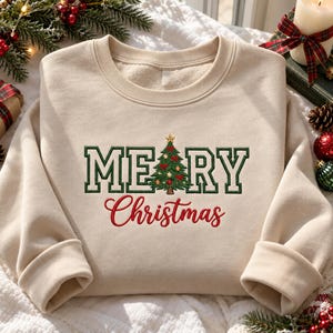 Könnte beinhalten: Ein beigefarbenes Sweatshirt mit Rundhalsausschnitt und einer Stickerei mit der Aufschrift MERRY CHRISTMAS in Dunkelgrün und Rot. Der Buchstabe A ist durch einen detaillierten grünen Weihnachtsbaum mit goldenen Kugeln, roten Schleifen und einem goldenen Stern ersetzt.