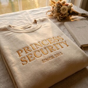 Może przedstawiać: Beżowa bluza z okrągłym dekoltem i złotym haftem na piersi. Napis brzmi PRINCESS SECURITY dużymi literami, a poniżej znajduje się mniejszy tekst SINCE 2013. Materiał ma gładkie wykończenie w jednolitym kolorze.