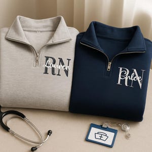 May include: Two quarter-zip sweatshirts, one light grey and one navy blue, embroidered with names and "RN". A stethoscope and a nurse badge are in the foreground. The sweatshirts have a high collar and a silver zip.