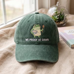 May include: A forest green baseball cap featuring an embroidered graphic of a frog wearing an apron, holding a wooden spoon and a bowl. Below the frog, the text WE PROOF AT DAWN is embroidered in white thread. The cap has a structured crown.