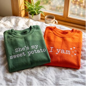Op de afbeelding: Een donkergroen sweatshirt met witte geborduurde tekst "She's my sweet potato" en een oranje sweatshirt met witte geborduurde tekst "I yam", vergezeld door verschillende kleine witte geborduurde hartjesomlijningen.