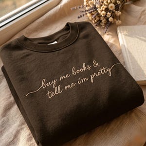 May include: A dark brown crewneck sweatshirt featuring light tan embroidered cursive text on the chest that reads, "buy me books & tell me i'm pretty." The garment has a ribbed collar and long sleeves, showcasing a simple and clean design.