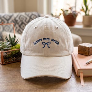 Puede incluir: Gorra de béisbol color crema con el texto bordado "future mrs. smith" en azul. Un lazo azul está bordado entre las palabras. La gorra tiene una visera desgastada y está sobre una superficie de madera.