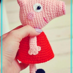 May include: A handmade crochet pig doll with a pink head, ears, and snout, a white eye, and a red dress. The doll has black feet and is held by a hand.