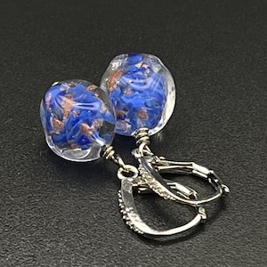 Può includere: Un paio di orecchini in vetro trasparente con inclusioni blu e color rame. Gli orecchini hanno chiusure a leva argentate. I globi di vetro sono rotondi e riempiti di elementi decorativi.