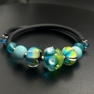 Puede incluir: Una pulsera con una banda negra y varias cuentas de vidrio de colores. Las cuentas son en tonos de azul, verde y amarillo, con detalles plateados. La pulsera tiene un diseño moderno y artístico.