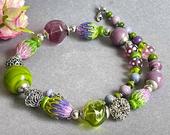 May include: A multi-strand necklace with green, purple, and white glass beads. The beads are various shapes and sizes, including flowers, polka dots, and swirls. The necklace is accented with silver metal beads and a silver clasp.