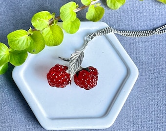 Op de afbeelding: Een zilveren ketting met een rode glazen frambozen hanger. De hanger heeft twee frambozen op een zilveren steel met een blad.
