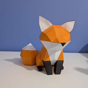 May include: A geometric paper fox sculpture in orange, white, and black. The fox has a triangular body, pointed ears, and a fluffy tail. The sculpture is sitting on a white surface against a blue background.