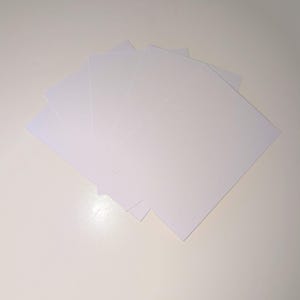Pode incluir: Um conjunto de folhas de papel retangulares brancas, ligeiramente abertas, mostrando as suas bordas e cantos. Estão dispostas sobre uma superfície branca, criando uma estética limpa e minimalista. As folhas estão em branco.