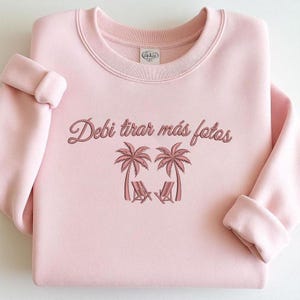 Puede incluir: Sudadera rosa claro con la frase "Debi tirar más fotos" en cursiva. Debajo, dos palmeras y dos sillas de playa. La sudadera tiene cuello redondo y mangas largas.