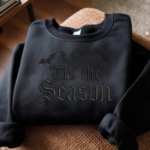 May include: Black sweatshirt with the phrase "Tis' the Season" in gothic lettering, accompanied by bat silhouettes. The long-sleeved crewneck top is made of a soft material.