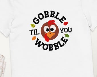 Może przedstawiać: Biała koszulka z okrągłą grafiką przedstawiającą kreskówkowego indyka i napisem "GOBBLE TIL YOU WOBBLE". Projekt zawiera jesienne liście w kolorze pomarańczowym i zielonym. Koszulka jest wykonana z miękkiego materiału.