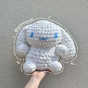 May include: A white crochet plush toy with blue embroidered features, including eyes and a mouth. The toy has large, floppy ears and is held by a hand. The image includes the text "@saffronsqueaks".
