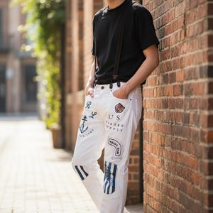 May include: A person wearing a black t-shirt and white jeans with nautical patches. The jeans have patches with anchors, the text "US", and "NAVAL DOCK". The person is wearing brown suspenders and white sneakers.
