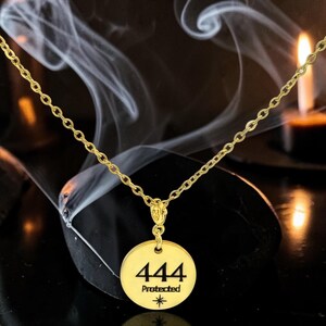 May include: A gold-toned necklace featuring a circular pendant with the number 444 engraved in black, followed by the word Protected and a small star symbol. The pendant hangs from a delicate gold-toned chain link, creating a simple jewelry piece.