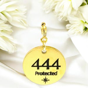 May include: A gold-colored circular charm featuring a lobster claw clasp. The front of the charm is engraved with the number 444, the word Protected, and a small star-like symbol. The surface of the metal is smooth and reflective.