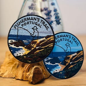 Pode incluir: Duas etiquetas redondas com o texto "Fisherman's Trail Portugal" e um gráfico de pescador. Uma etiqueta é bordada com cores azul, castanho e amarelo. A outra é um autocolante com um design azul e castanho.