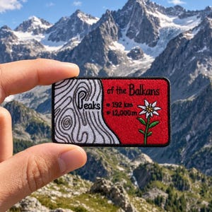 May include: A rectangular embroidered patch featuring a white topographic map design on the left and a red background on the right. The text reads Peaks of the Balkans, 192 km, and 12,000 m, accompanied by an embroidered white edelweiss flower.